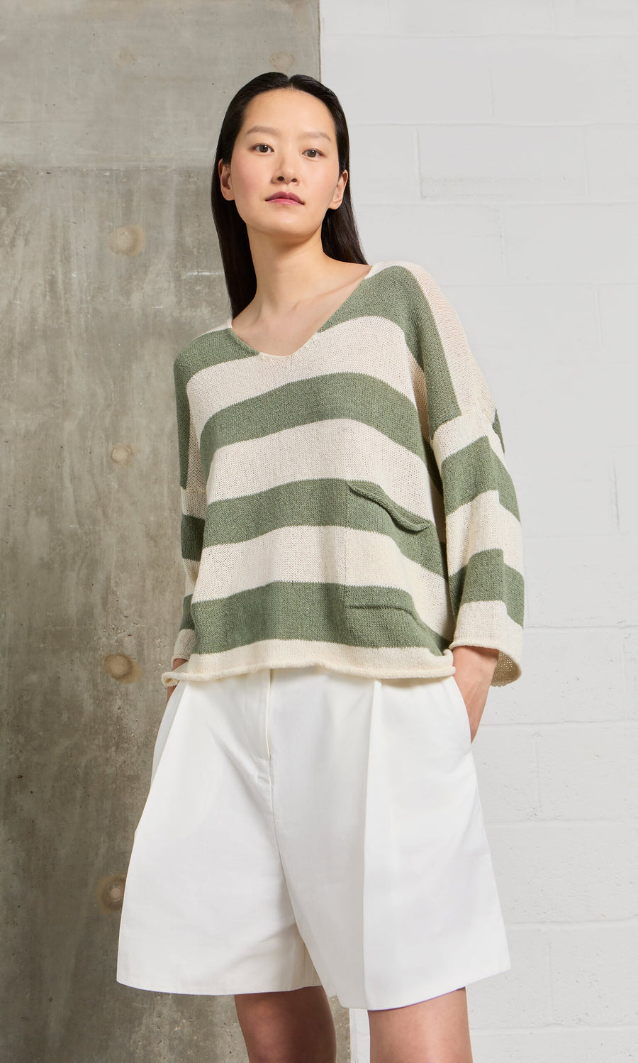 Plümo- Woman models the Hagen Stripe Jumper, a relaxed fit sage and white V-neck sweater, with white shorts.