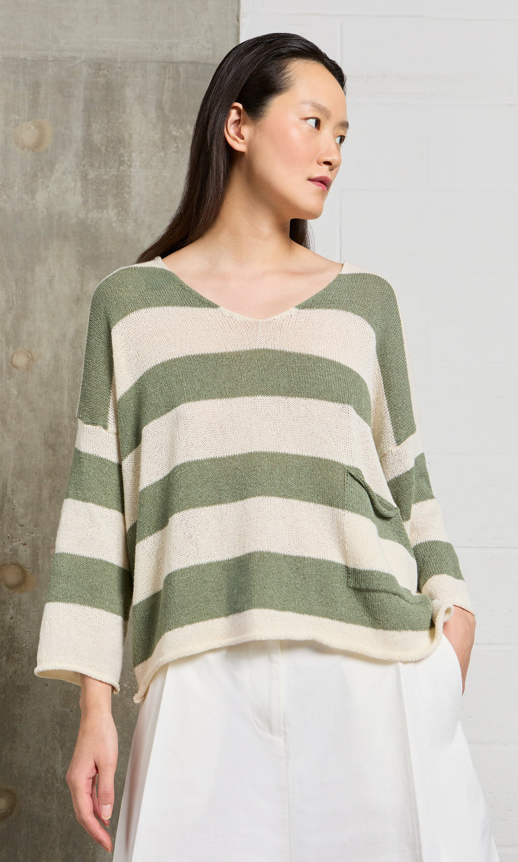 Plümo- Woman in the Hagen Stripe Jumper, a relaxed fit V-neck with sage and white stripes, paired with white pants.