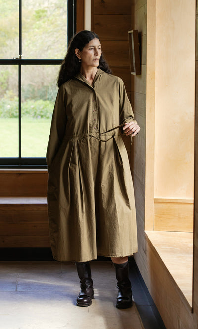 Plümo  Woman in a Hazel Dress and boots stands by a window, looking out, hand resting on a wooden ledge.