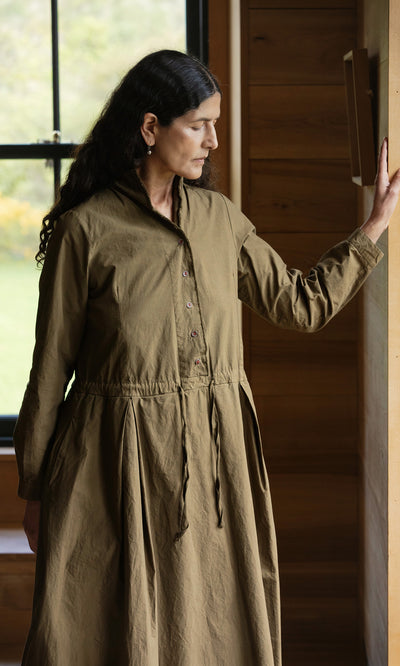 Plümo  Woman in a hand-dyed cotton Hazel Dress stands by a window, touching a wooden wall and looking down.