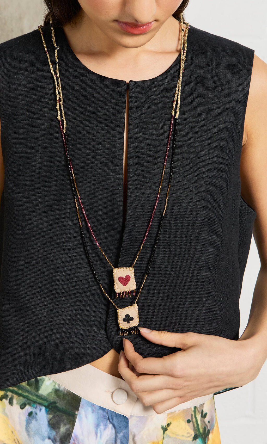 Plümo- A woman wears the Hearts & Clubs Necklace, a sleeveless black top, and floral pants.