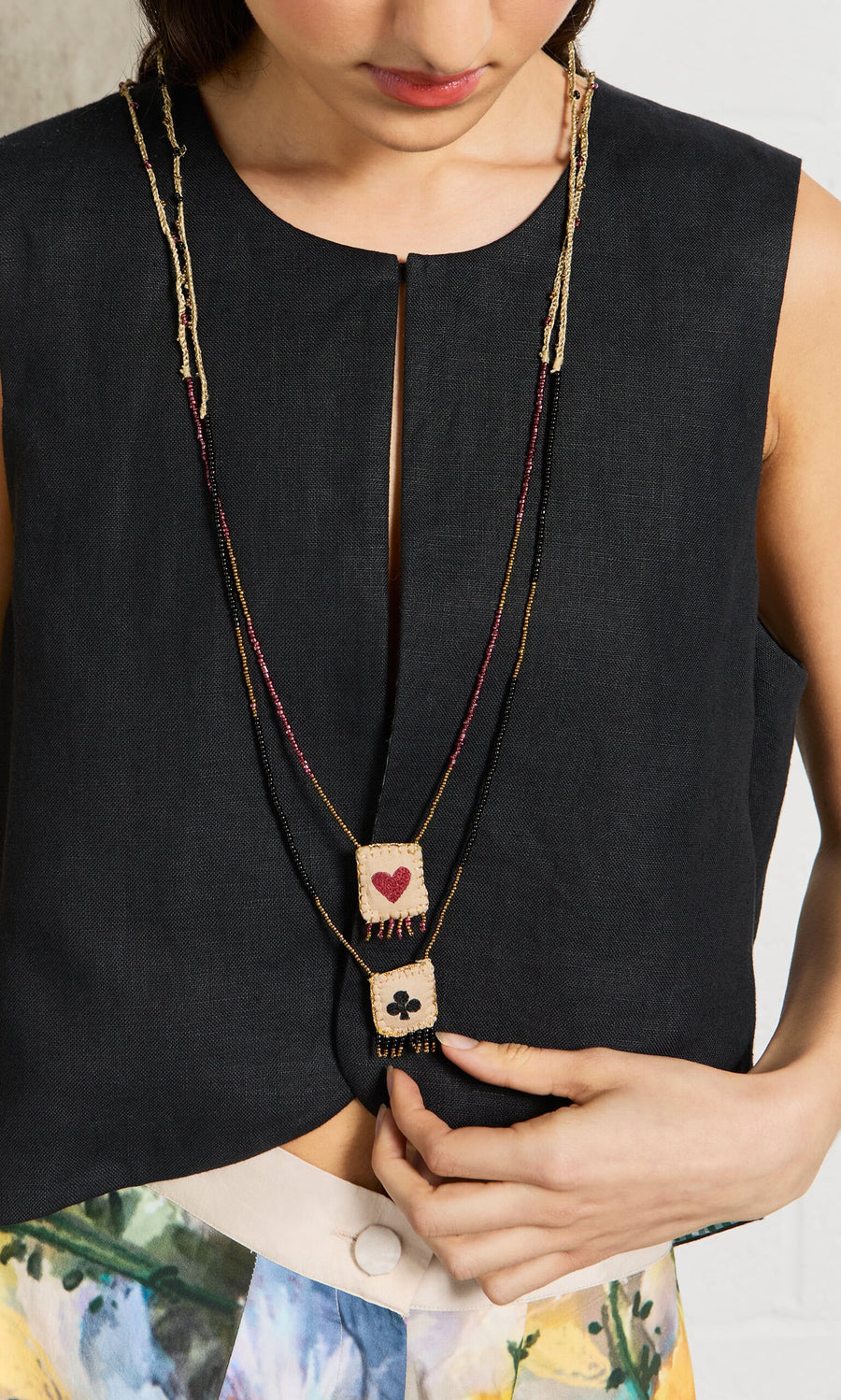 Plümo- A woman wears the Hearts & Clubs Necklace, a sleeveless black top, and floral pants.