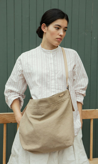 Plümo  Woman in a striped shirt with the Paloma Bag, featuring a crossbody strap, stands by a green wall, looking aside.