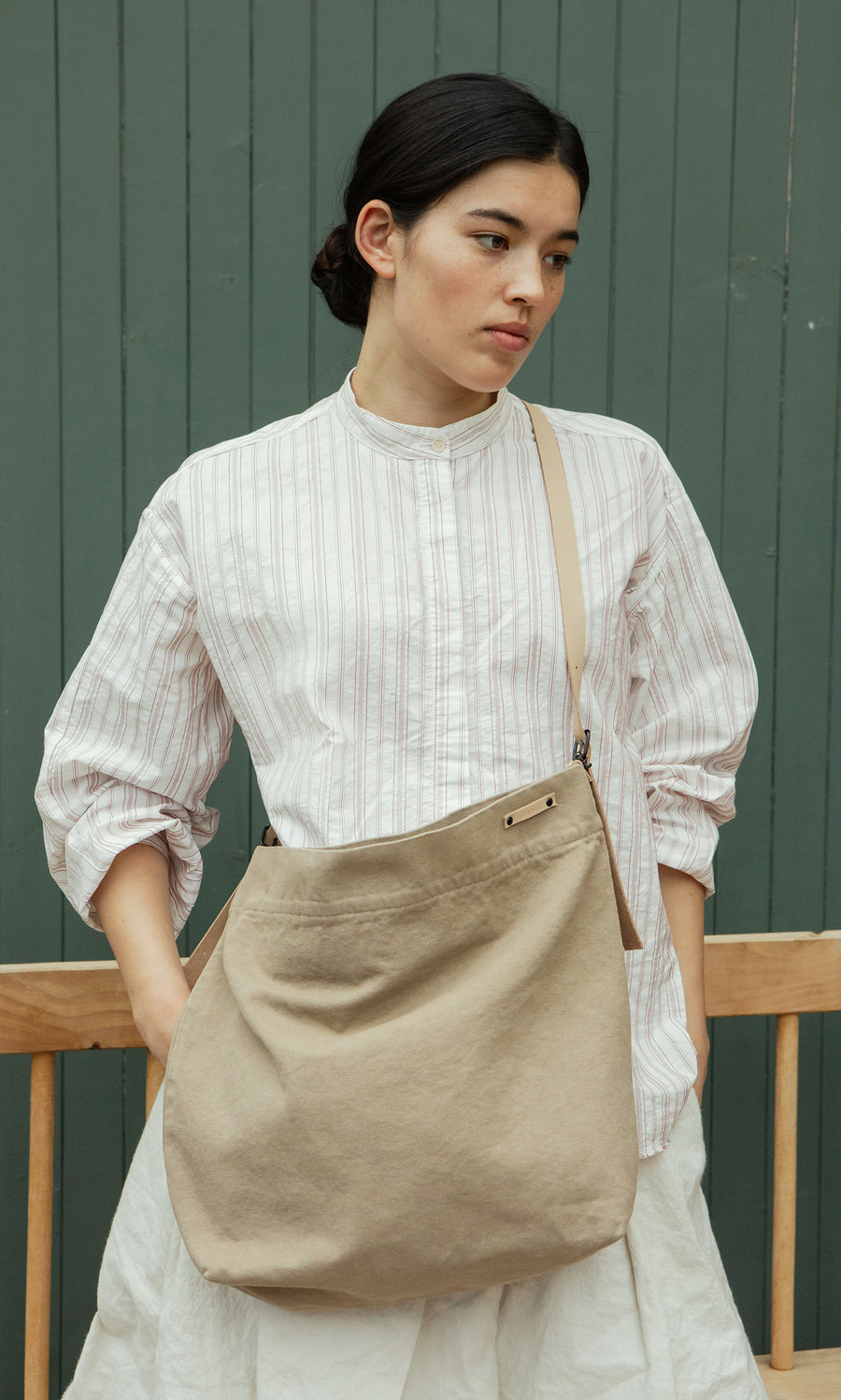 Plümo  Woman in a striped shirt with the Paloma Bag, featuring a crossbody strap, stands by a green wall, looking aside.