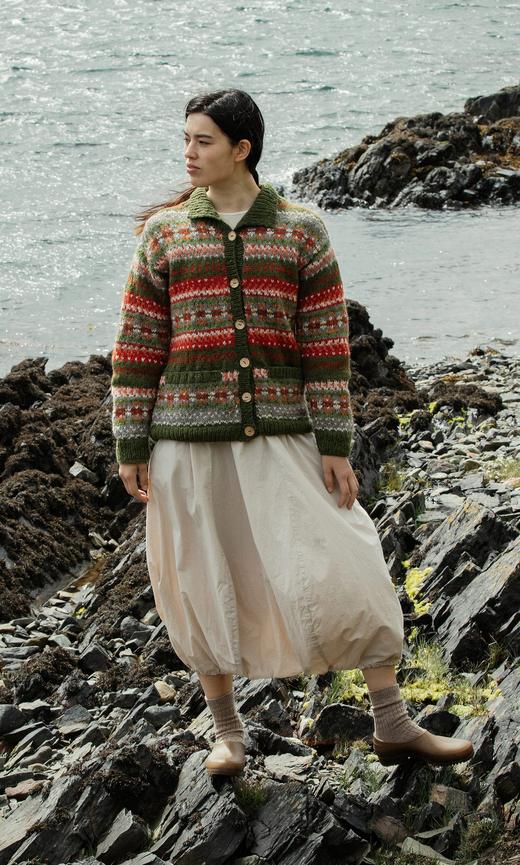 Plümo  Woman in the Hebrides Cardigan and a cream skirt stands on a rocky shore by the water, looking to the side.