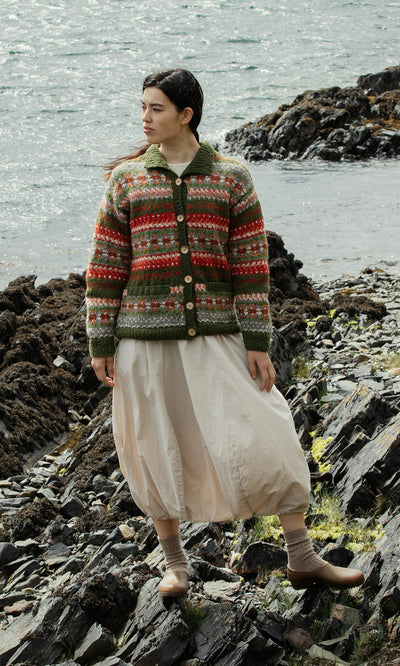 Plümo  Woman in the Hebrides Cardigan and a cream skirt stands on a rocky shore by the water, looking to the side.