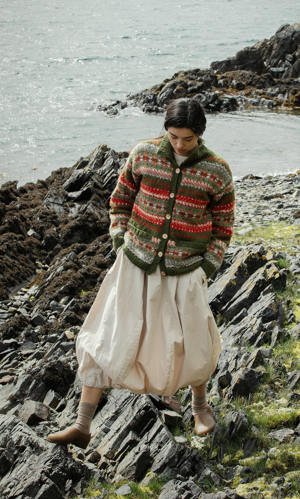 Plümo  Someone in the Hebrides Cardigan and cream skirt walks on rocky ground by the water.