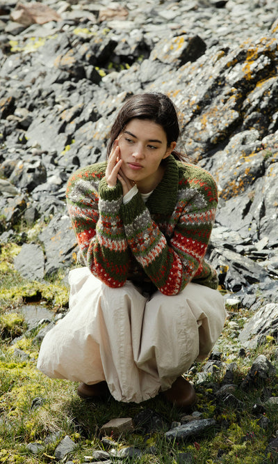 Plümo  Someone in the Hebrides Cardigan and a cream skirt squats on rocks, resting their chin thoughtfully on their hand.