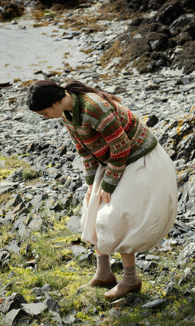 Plümo  Woman in a Hebrides Cardigan and white skirt bends over on a rocky, grassy shore by the water.