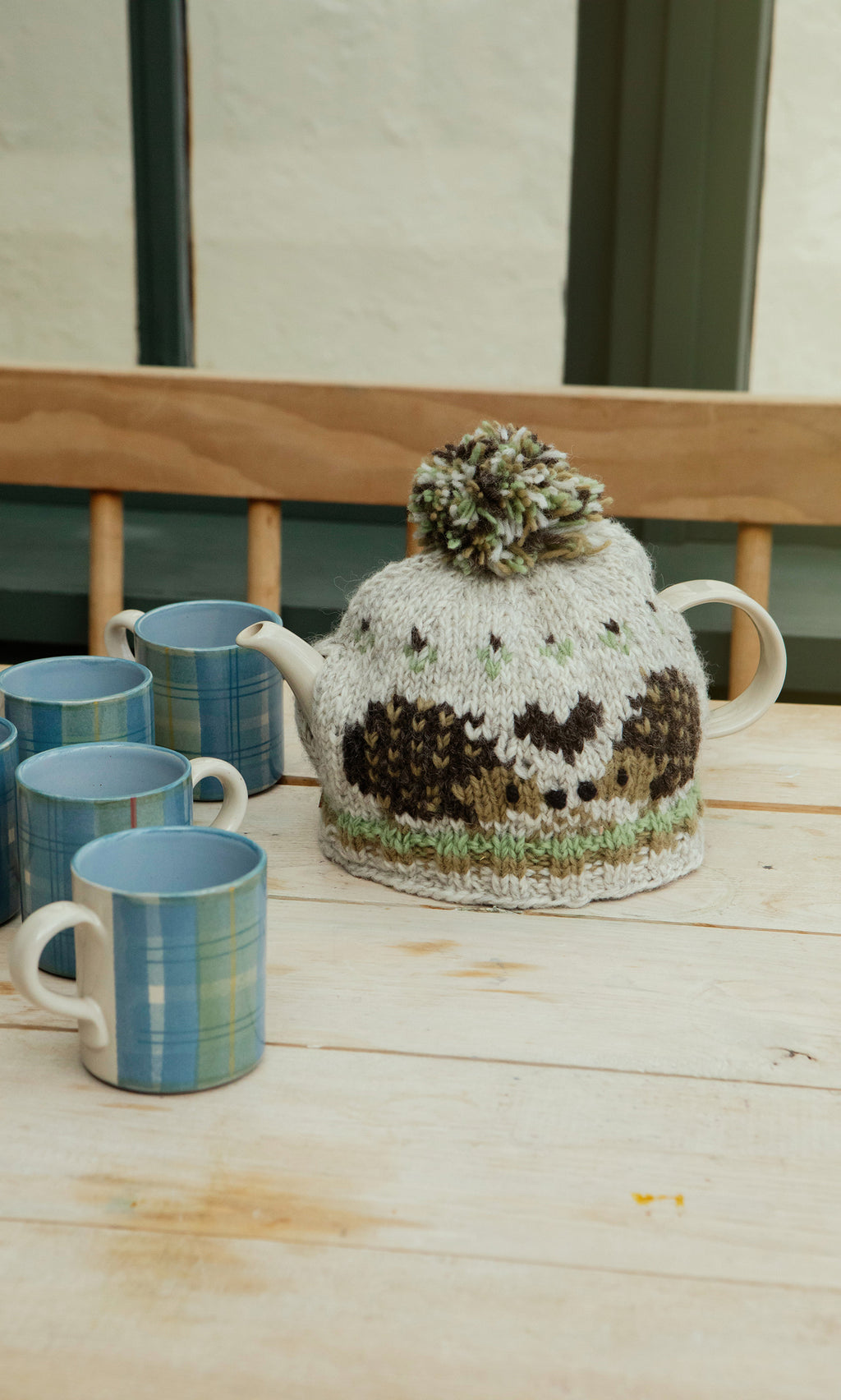 Plümo  A fairtrade teapot with a Hedgehog Tea Cosy and five blue plaid mugs sits on a light wood table near a window.