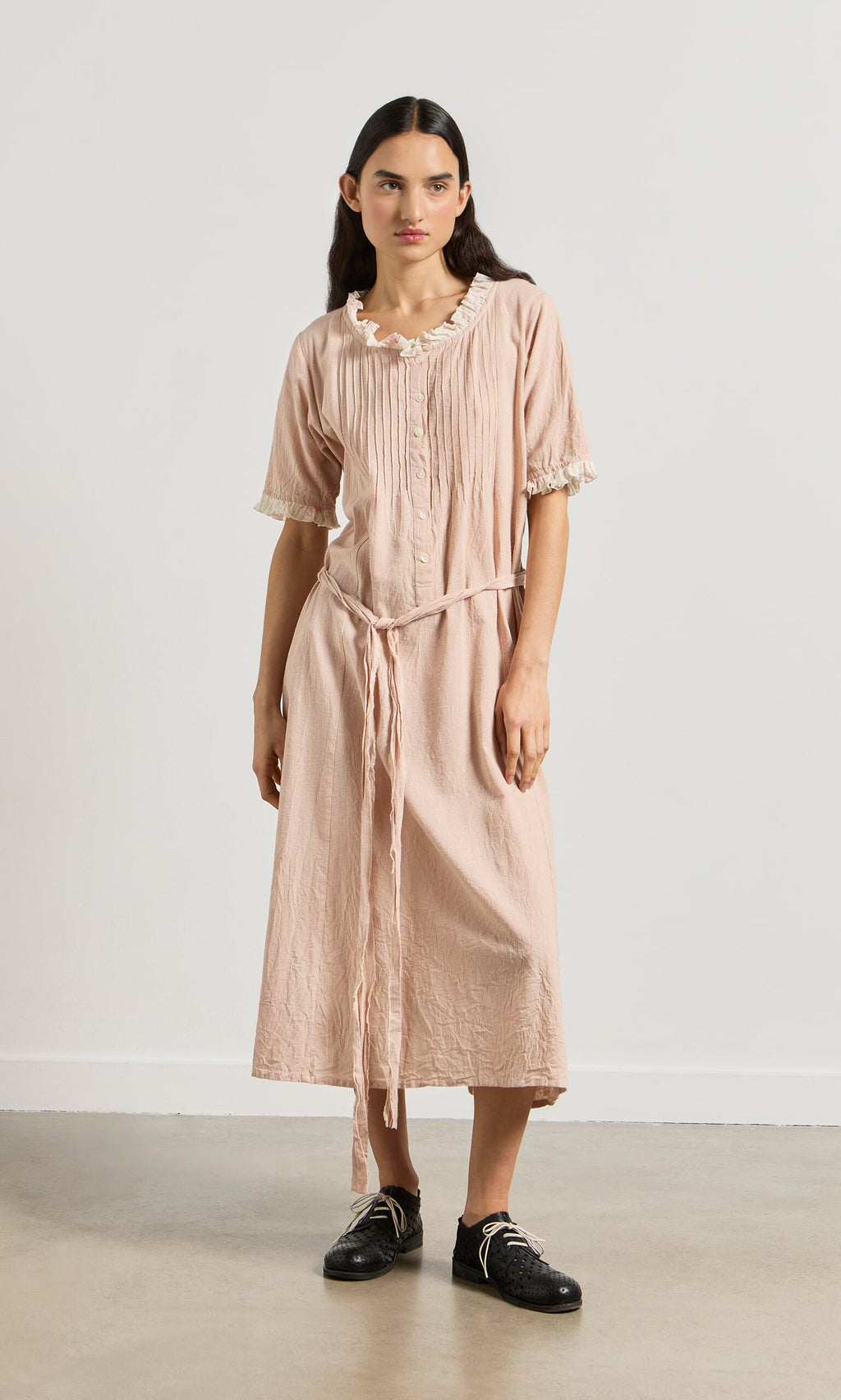Plümo- Woman wearing the Heide Dress in light pink with short sleeves and black shoes, standing against a plain background.
