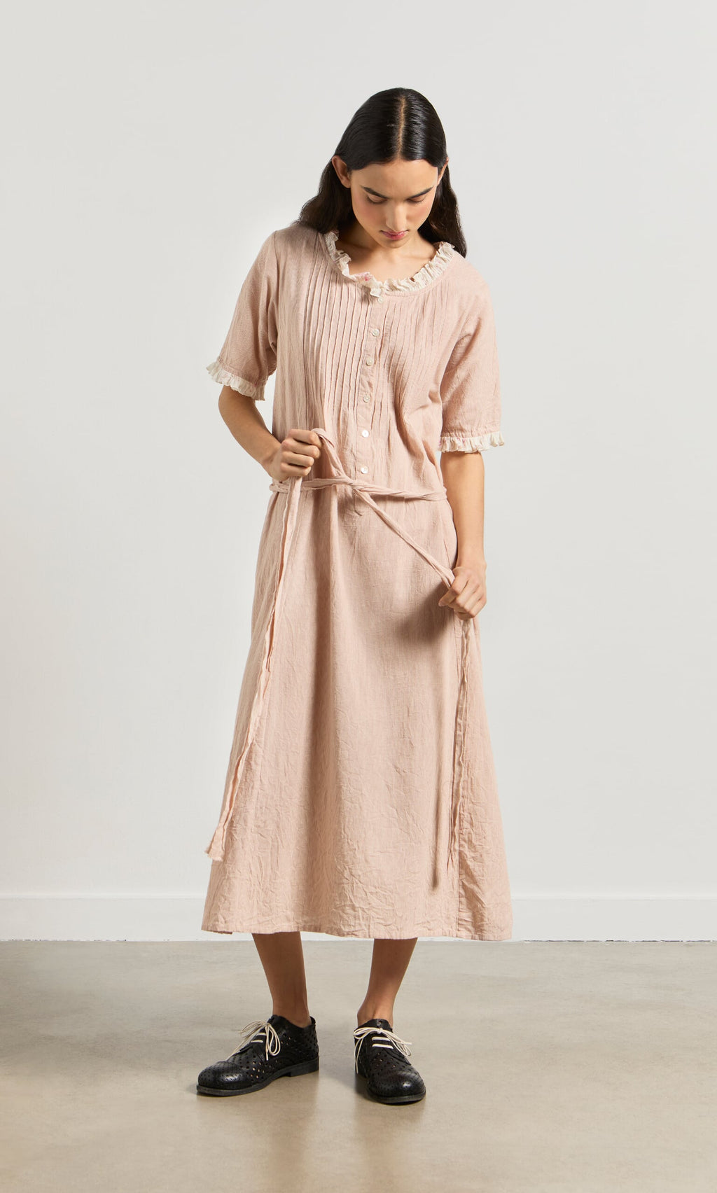 Plümo- Woman in the Heide Dress with lace trim, tying the waist belt and looking down, standing indoors.