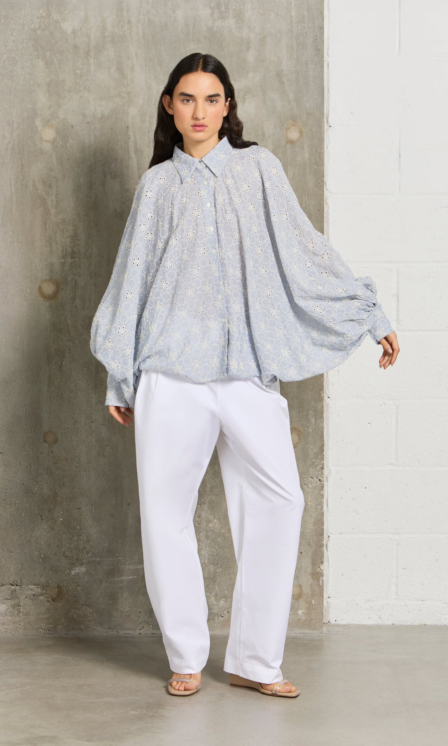 Plümo- A woman in the Hortence Blouse, a loose light blue top, and white pants stands indoors by a concrete and brick wall.