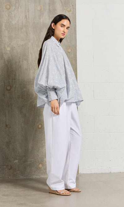 Plümo- A woman stands sideways in a Hortence Blouse with puffy sleeves and wide white trousers in a modern setting.