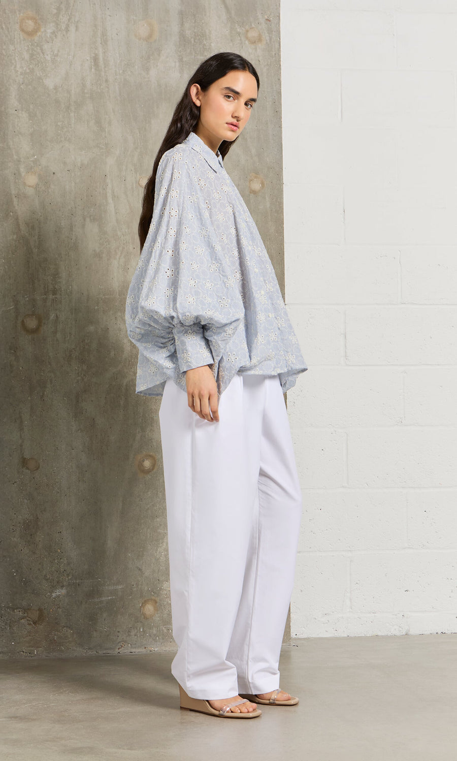 Plümo- A woman stands sideways in a Hortence Blouse with puffy sleeves and wide white trousers in a modern setting.