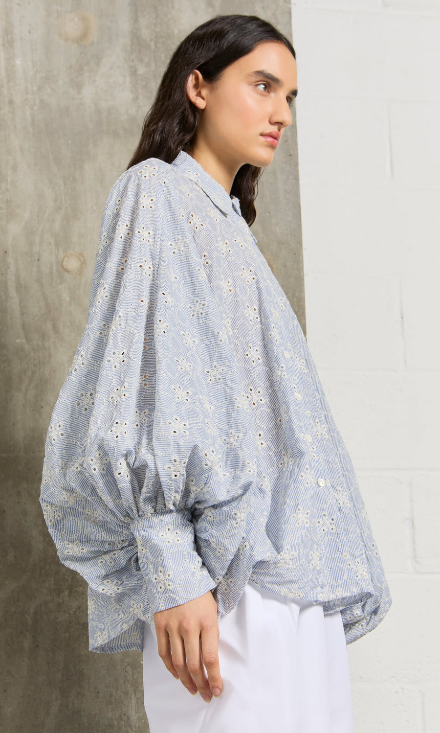 Plümo- A woman wears the Hortence Blouse, a loose blue broderie anglaise shirt with puffed sleeves, against a white wall.