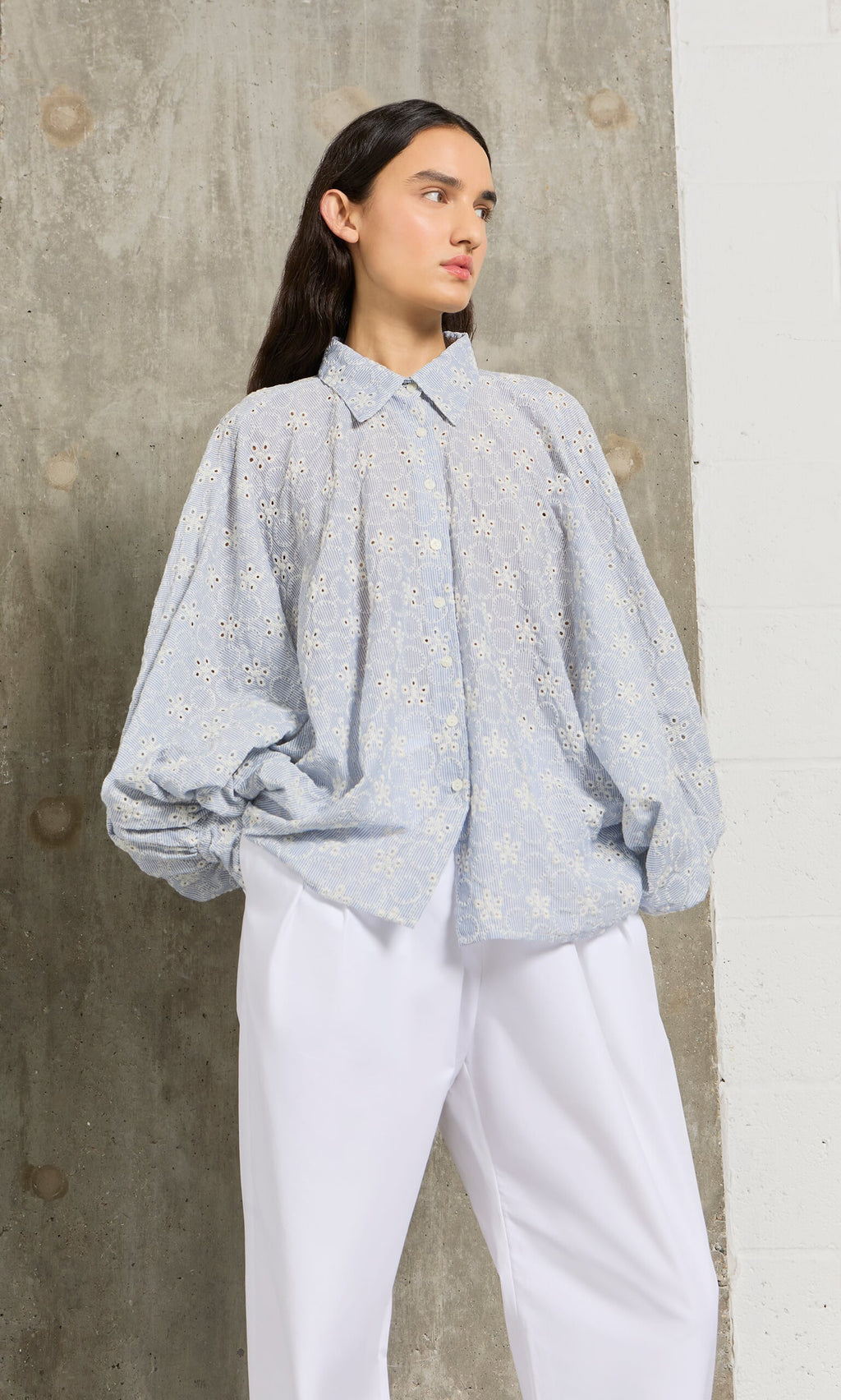 Plümo- Woman in the Hortence Blouse, a light blue broderie anglaise shirt, with white pants, stands against a concrete wall.
