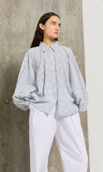 Plümo- Woman in the Hortence Blouse, a light blue broderie anglaise shirt, with white pants, stands against a concrete wall.