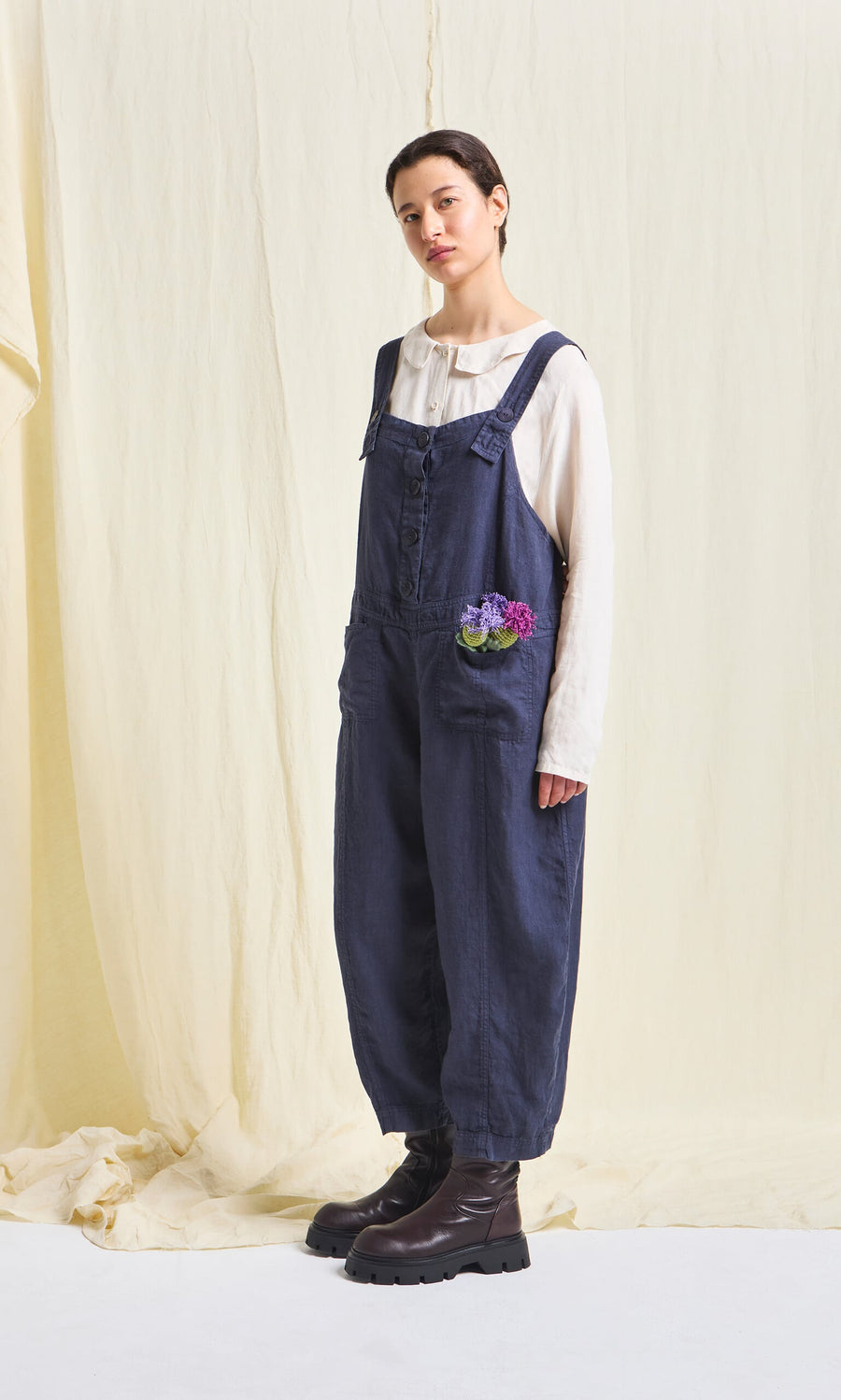 Plümo- Person in Hosie Linen Jumpsuit and white shirt stands on a light backdrop, flowers peeking from the pocket.