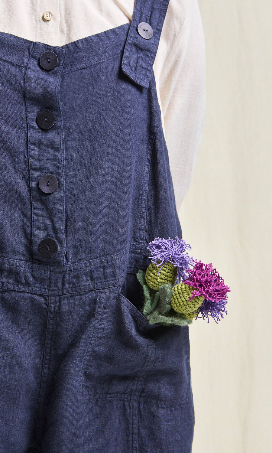 Plümo- Person in a white shirt and the Hosie Linen Jumpsuit with purple and green flowers in the front pocket.