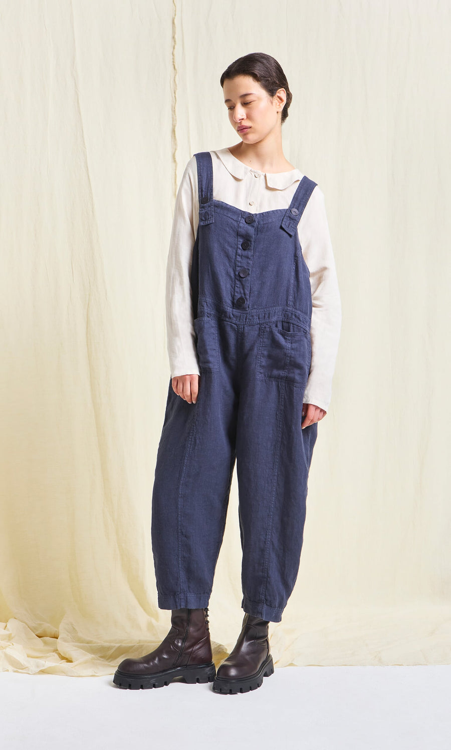 Plümo- Woman wears a Hosie Linen Jumpsuit in dark indigo with boots, standing against a light fabric backdrop.