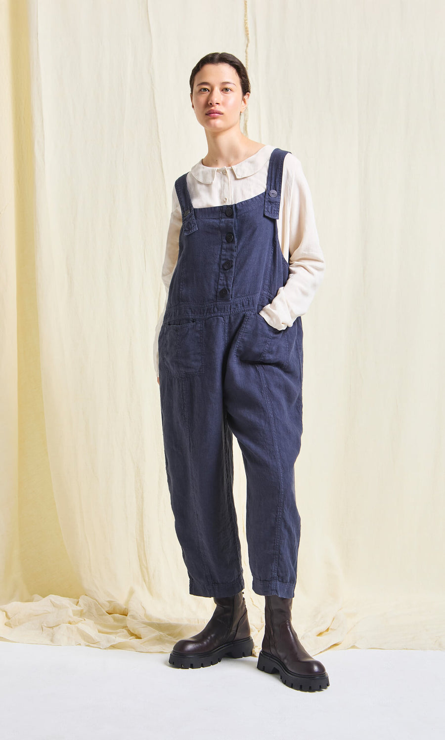 Plümo- Person standing with hands in pockets, wearing the Hosie Linen Jumpsuit, white blouse, and dark boots against a light background.