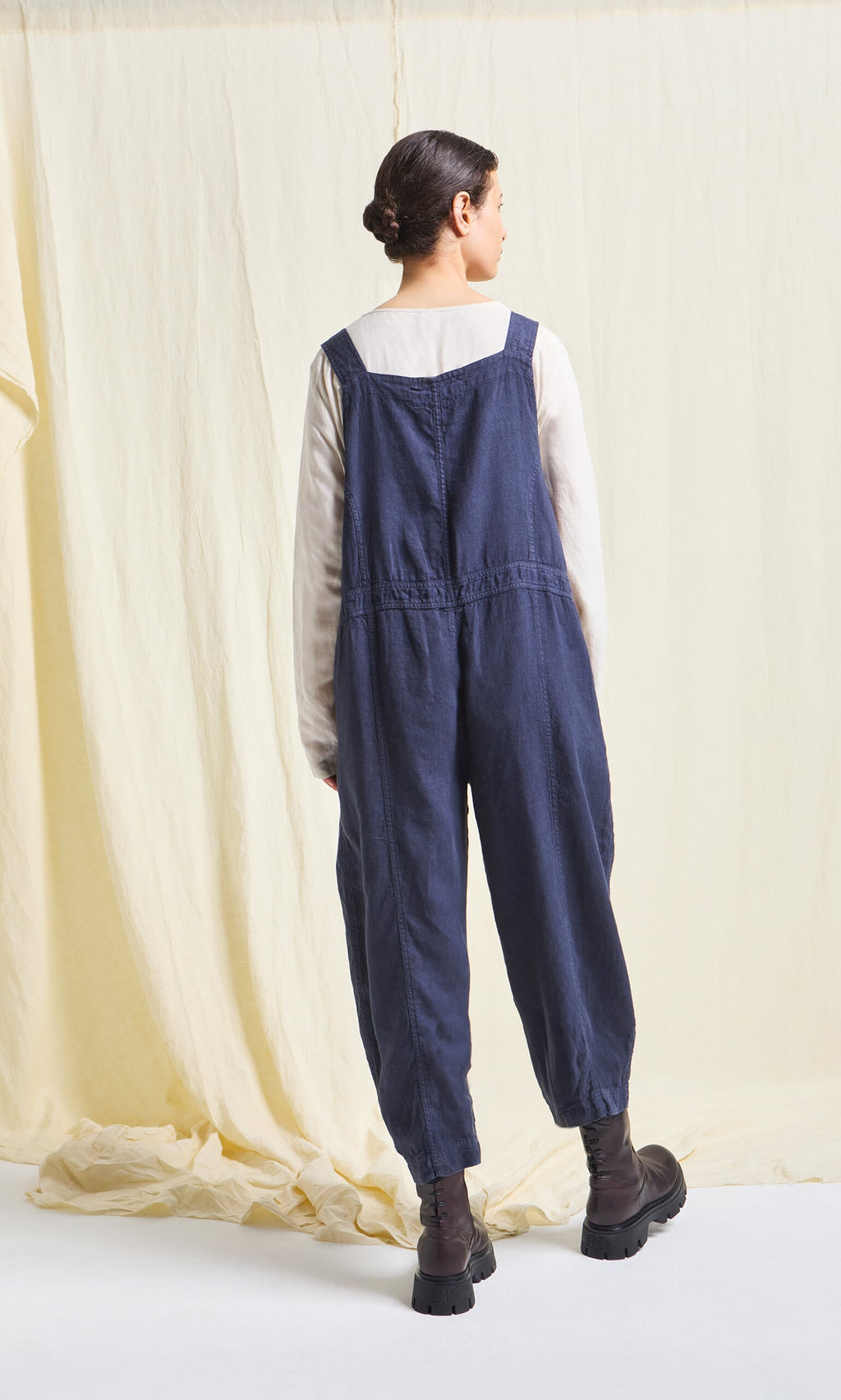 Plümo- Woman stands with back turned in an oversized cream top and Hosie Linen Jumpsuit, against a light fabric backdrop.