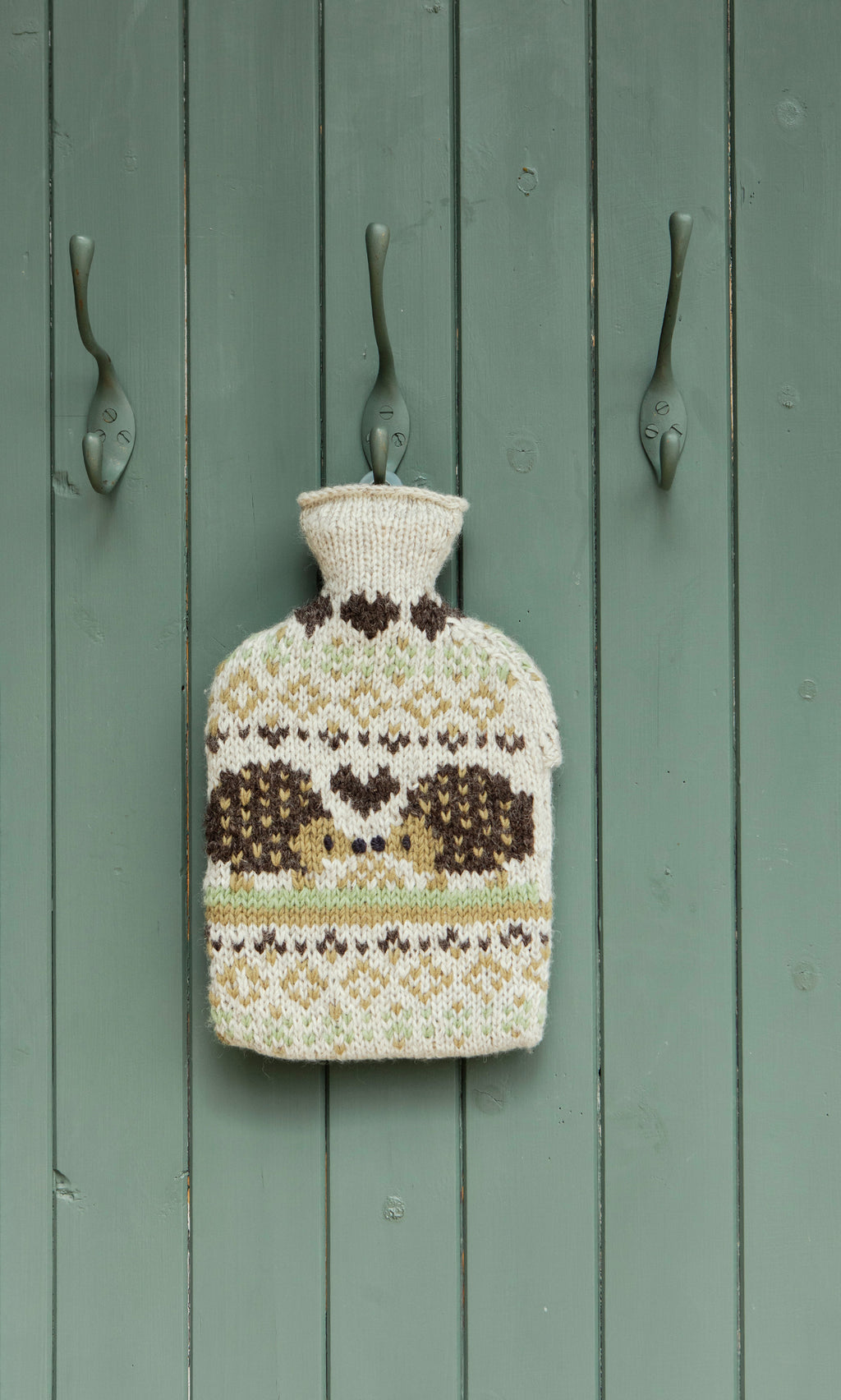 Plümo  The Hedgehog HWB hangs on a green door between three metal hooks, showcasing its hand-knitted patterned design.