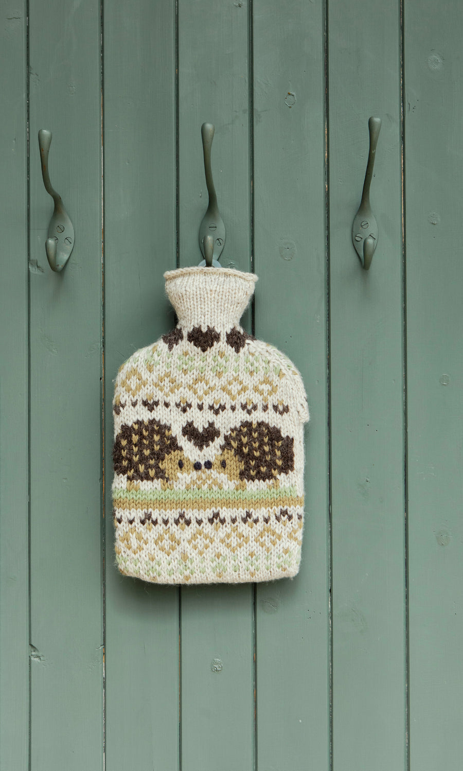 Plümo  The Hedgehog HWB hangs on a green door between three metal hooks, showcasing its hand-knitted patterned design.