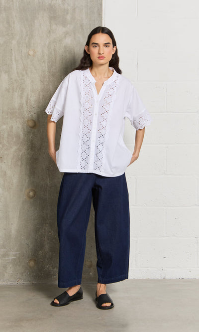 Plümo- Woman in the Hurlingham Blouse, wide-leg dark jeans, and black sandals stands by a concrete and white wall.