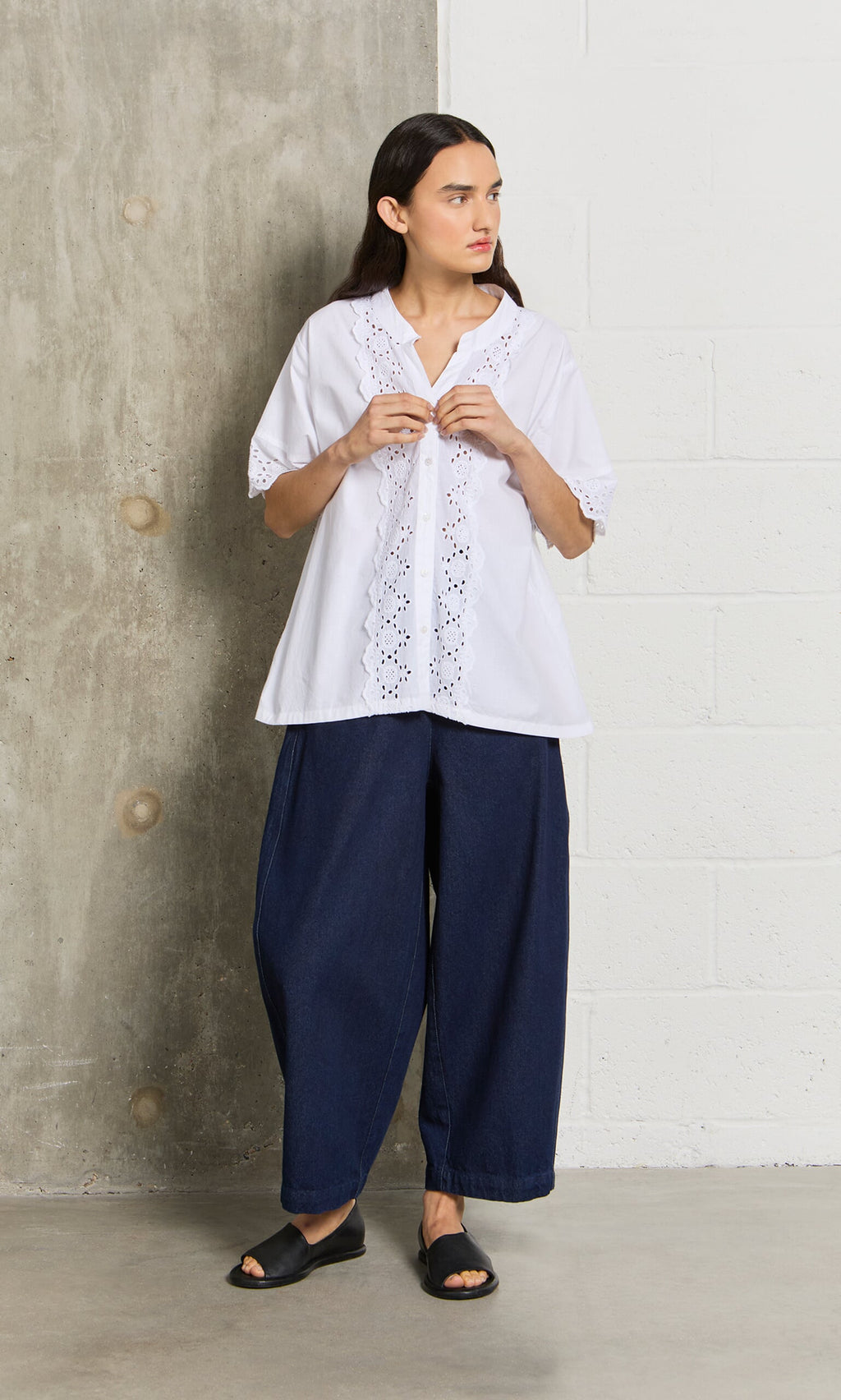 Plümo- Woman standing indoors in the Hurlingham Blouse, wide dark blue pants, and black sandals against a concrete wall.