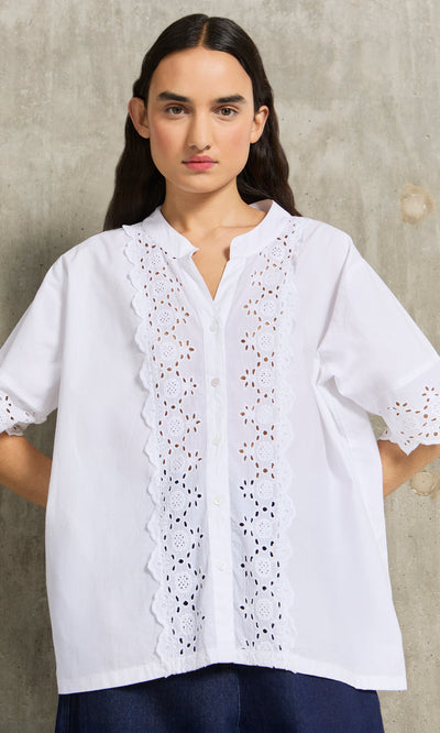 Plümo- Woman wears the Hurlingham Blouse, a white shirt with eyelet embroidery, standing against a concrete wall.