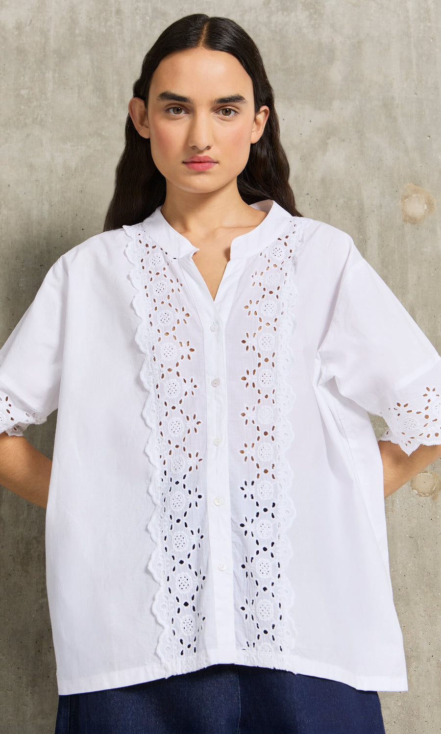 Plümo- Woman wears the Hurlingham Blouse, a white shirt with eyelet embroidery, standing against a concrete wall.