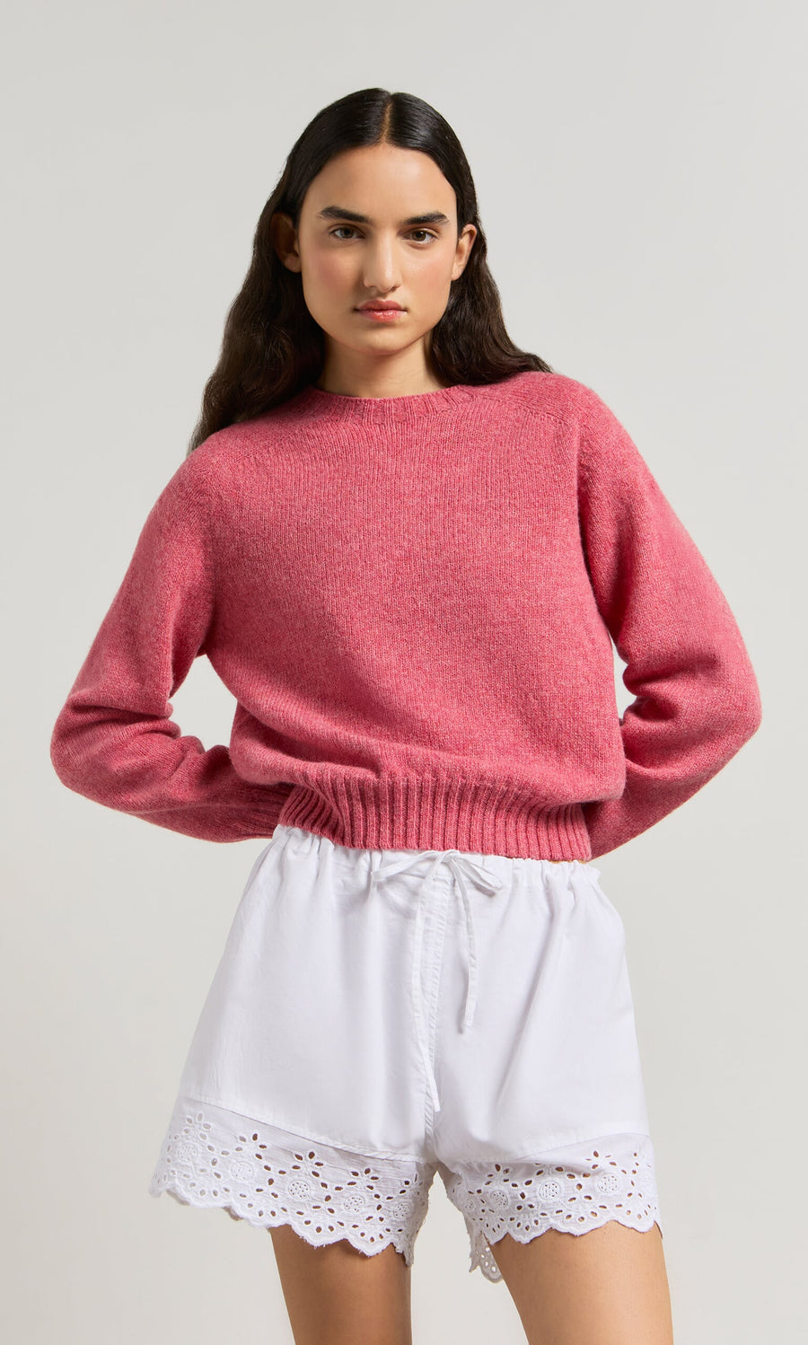 Plümo- Woman in a pink sweater and Hurlingham Shorts, standing with hands on hips against a plain background.