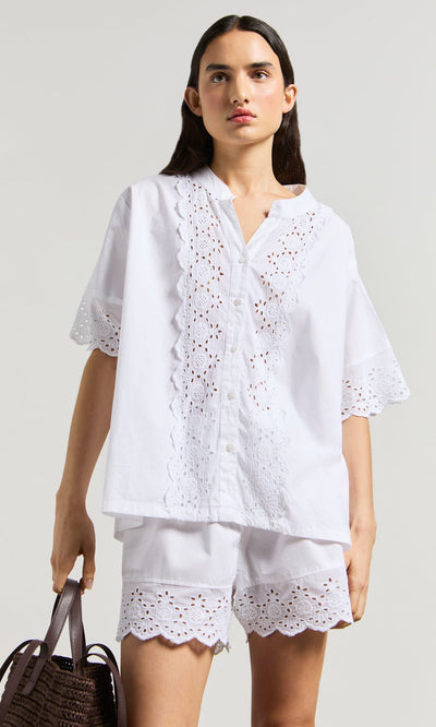 Plümo- Woman wears the Hurlingham Blouse and matching shorts, holding a brown woven bag against a plain background.