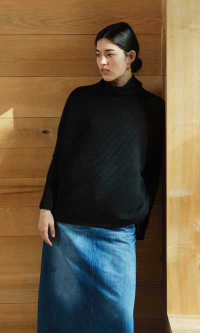 Plümo  Woman in the Jack Rollneck - Black Coal sweater leans against a wooden wall, looking to the side.