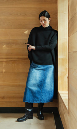 Plümo  Woman in Jack Rollneck - Black Coal sweater, denim skirt, and boots stands by a wooden wall, looking down.
