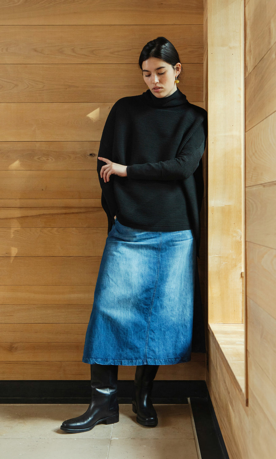 Plümo  Woman in Jack Rollneck - Black Coal sweater, denim skirt, and boots stands by a wooden wall, looking down.