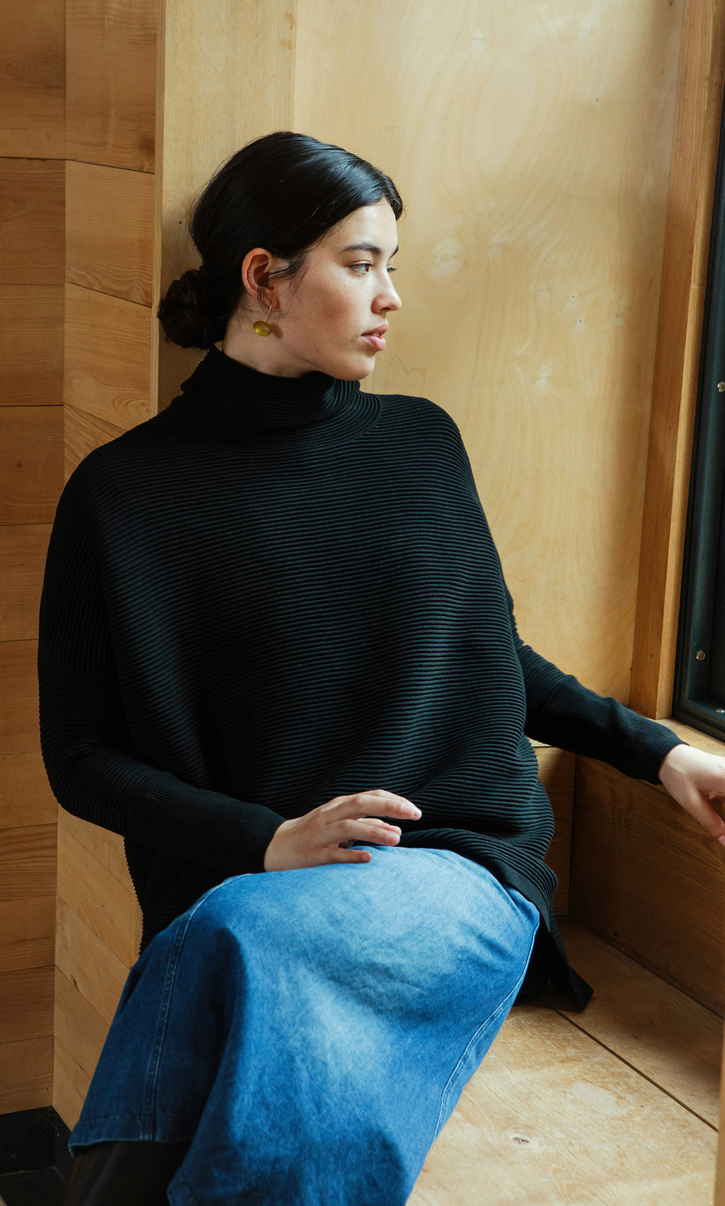 Plümo  Woman wears a Jack Rollneck in Black Coal and a blue skirt, sitting by a wooden wall and looking out the window.
