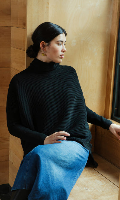 Plümo  Woman wears a Jack Rollneck in Black Coal and a blue skirt, sitting by a wooden wall and looking out the window.