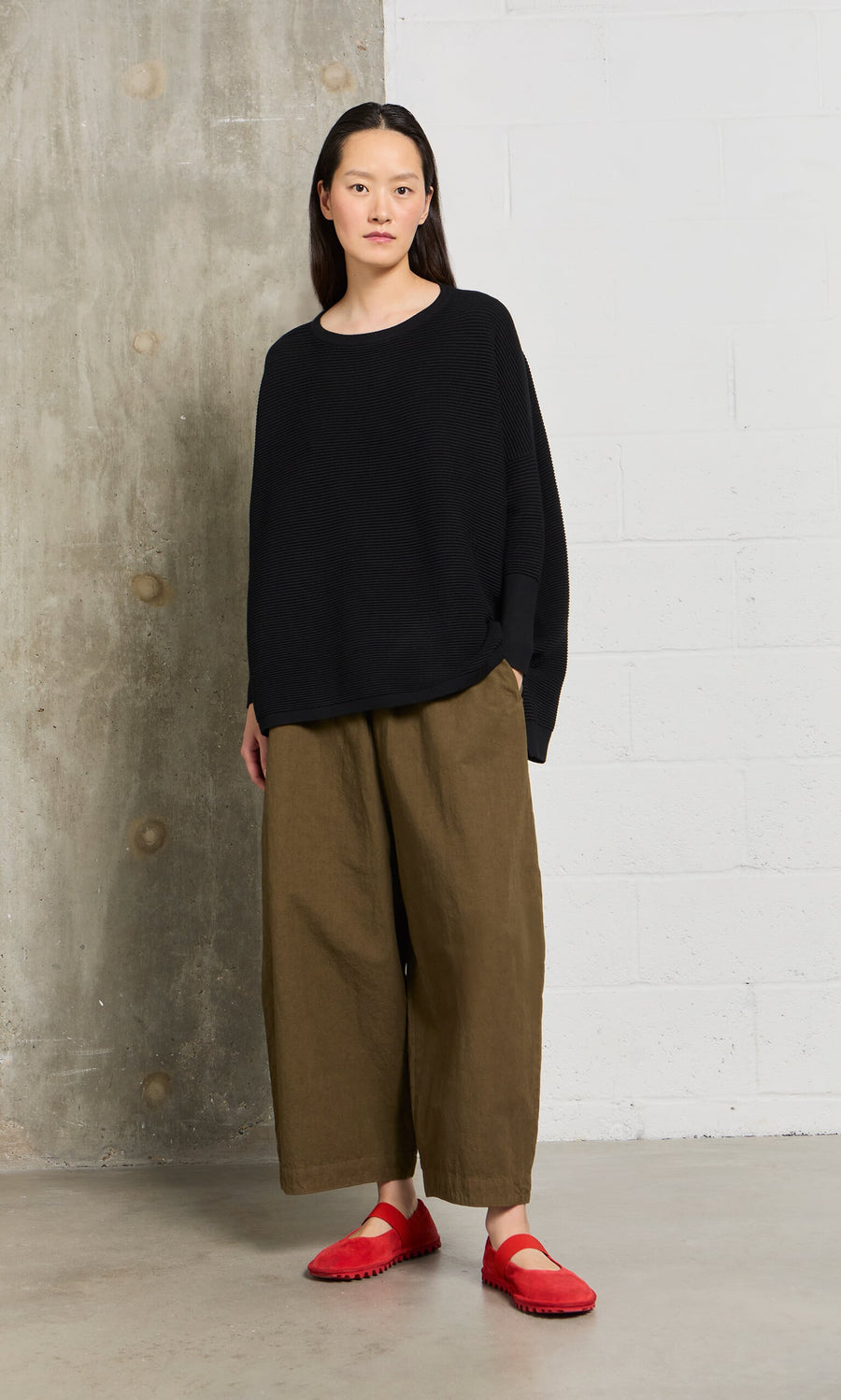 Plümo- A woman is indoors wearing the Jack Sweater in black, featuring dropped shoulders, with wide brown pants and red shoes.