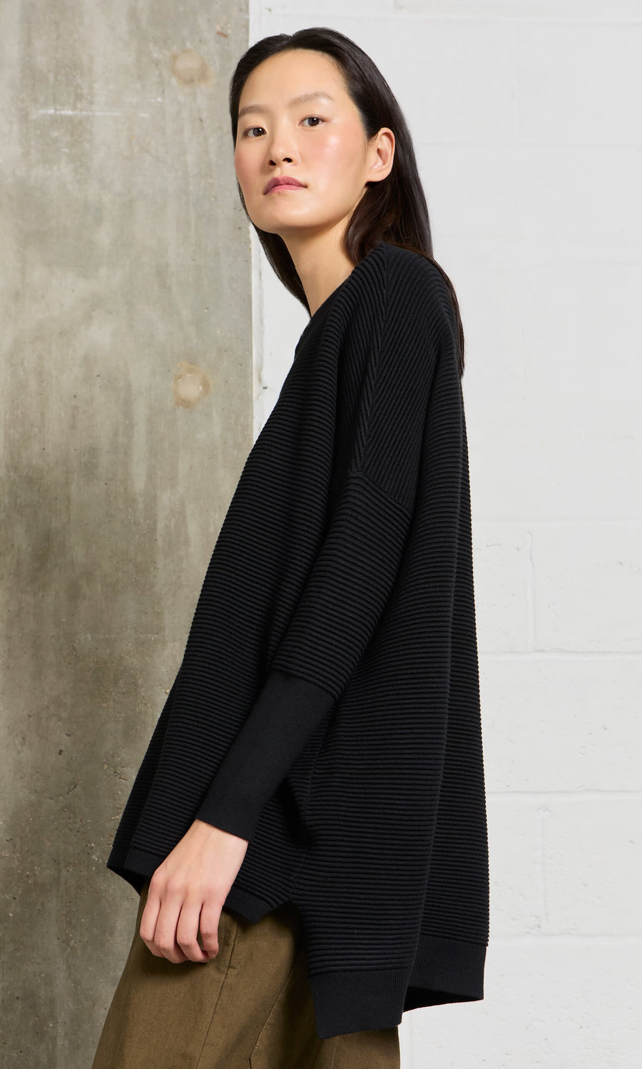 Plümo- Woman wearing the Jack Sweater – Black with dropped shoulders and brown pants, standing sideways by a brick wall.