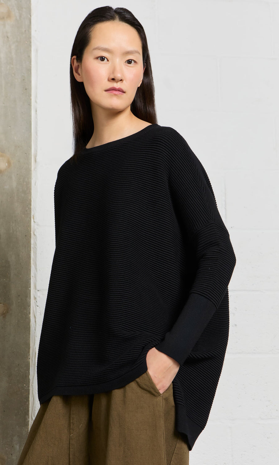 Plümo A person wears the Jack Sweater - Black, an oversized style with dropped shoulders, paired with brown pants.