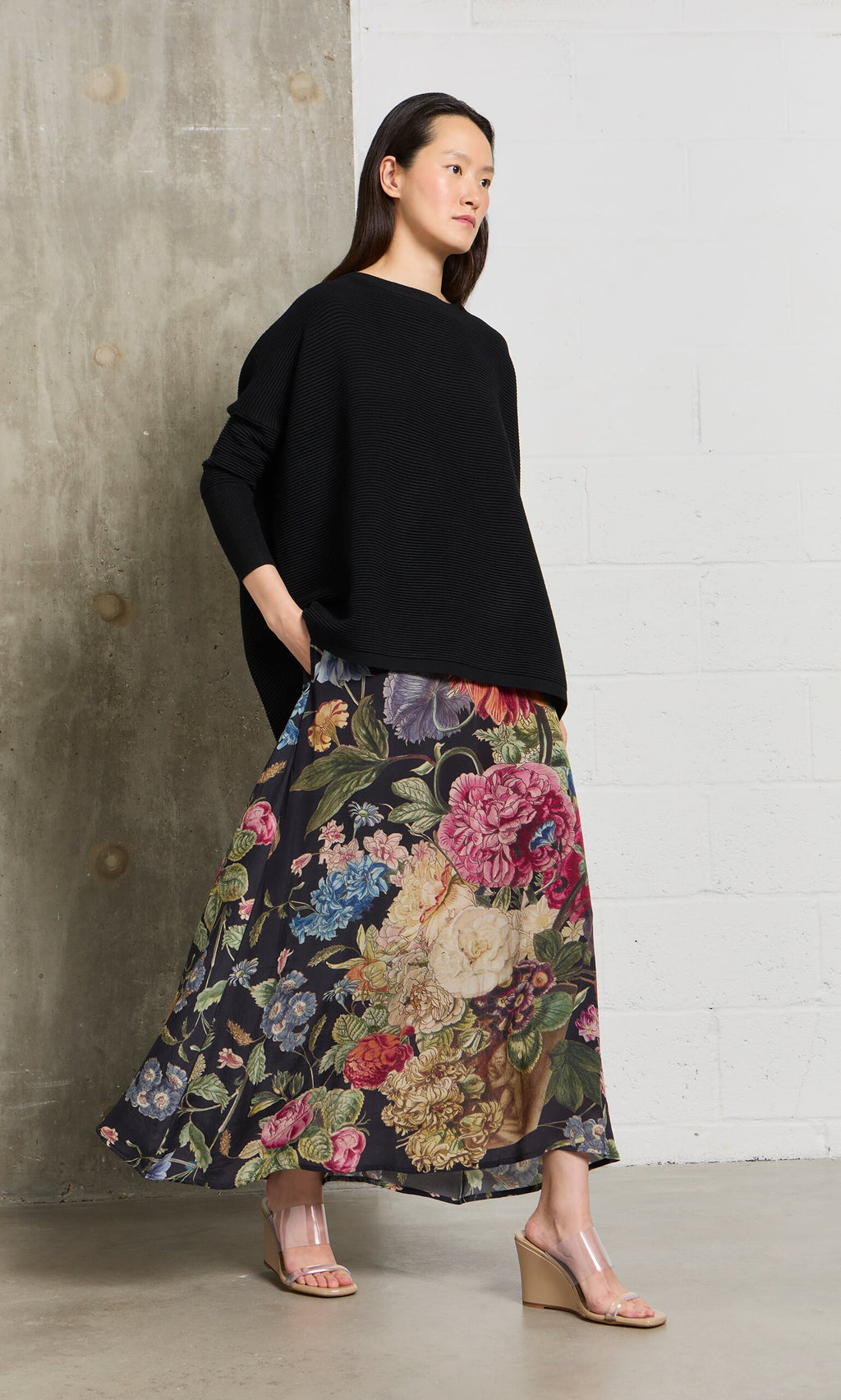 Plümo- Woman in the Jack Sweater - Black with dropped shoulders and a long floral skirt stands by a concrete and white wall.