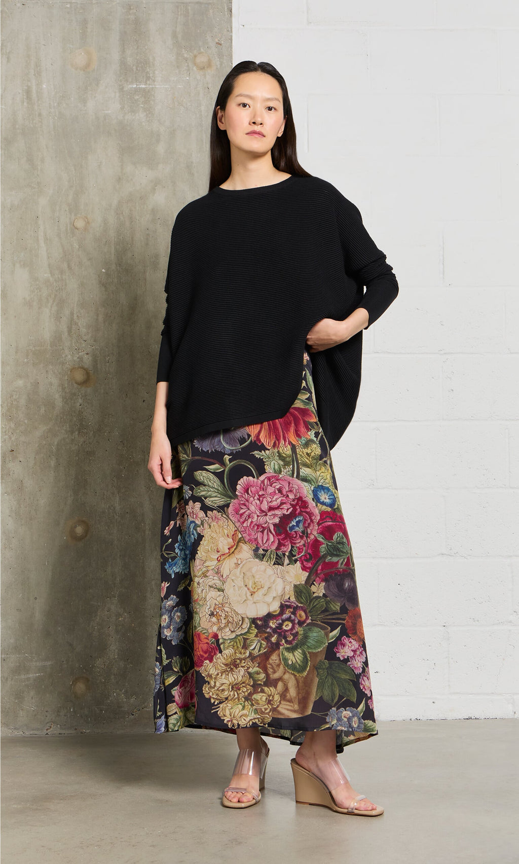 Plümo- Woman in a loose black sweater and the Auricula Skirt with wedge sandals, standing indoors against a concrete wall.