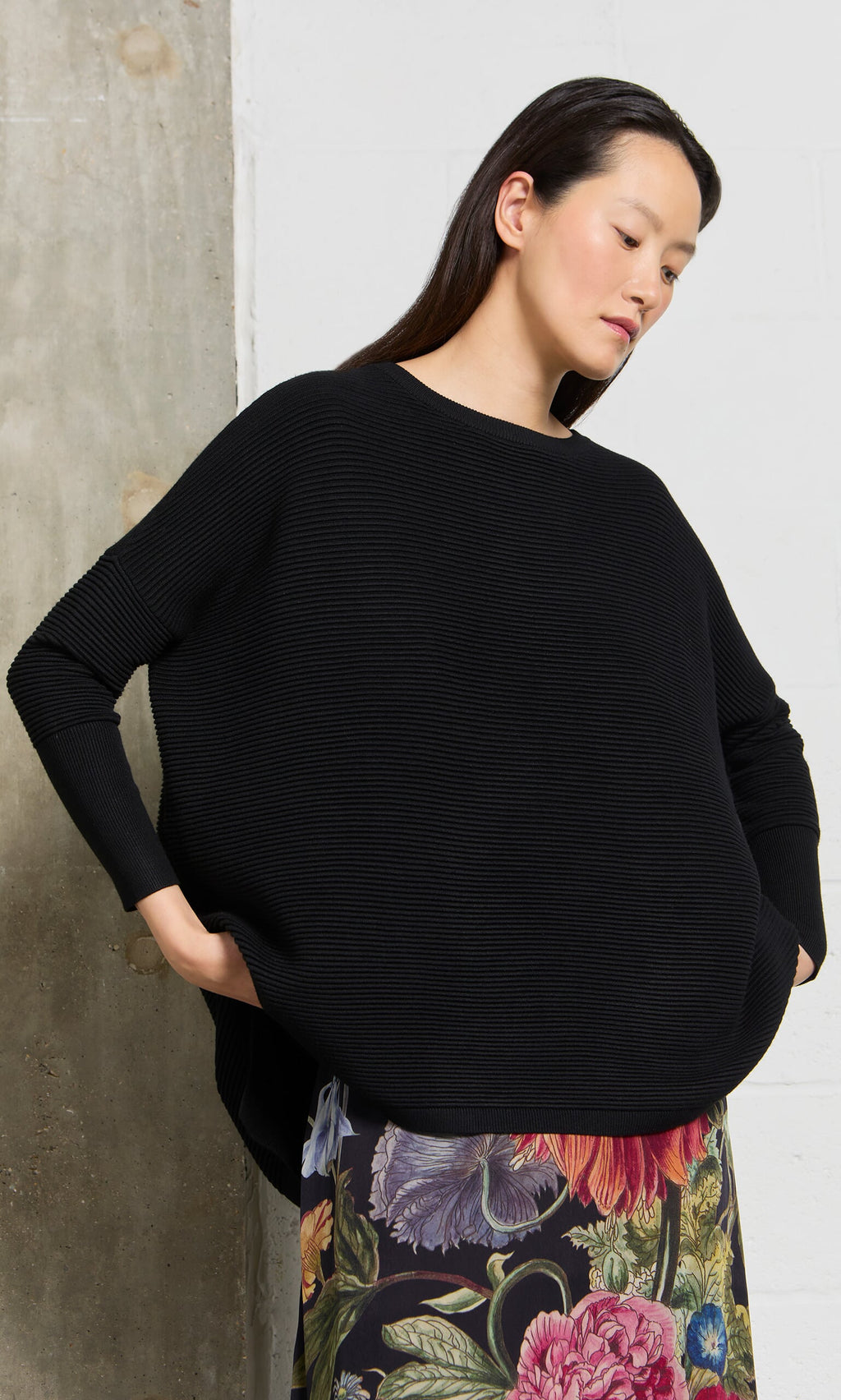 Plümo- Woman wearing the Jack Sweater - Black, oversized with dropped shoulders, floral skirt, hands in pockets.
