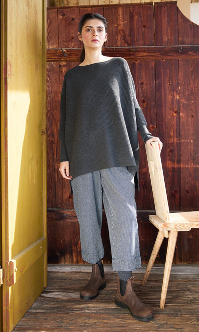 Plümo- Woman in Jack Sweater - Graphite, striped pants, and brown boots stands by a wooden chair and yellow door.