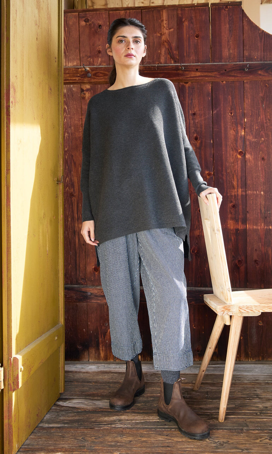 Plümo- Woman in Jack Sweater - Graphite, striped pants, and brown boots stands by a wooden chair and yellow door.