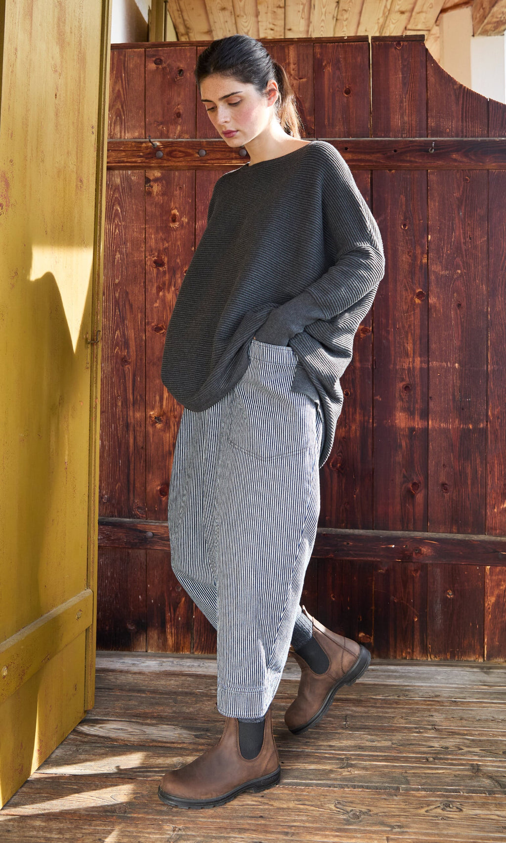 Plümo- A woman in the Jack Sweater - Graphite stands on a wooden porch, looking down.
