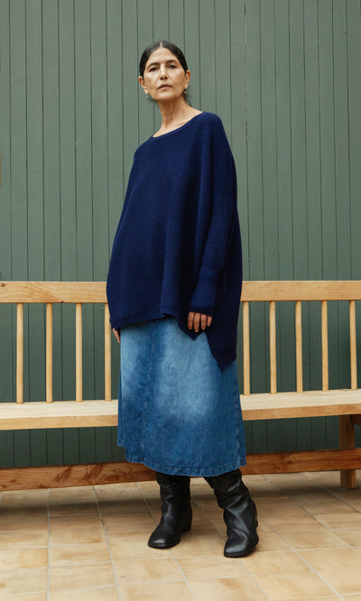 Plümo  Woman in the Jack Sweater - Navy, denim skirt, and boots stands by a wooden bench and green wall.